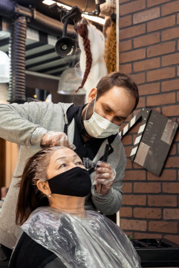 Bearded man in protective mask applying hair dye for aged female in modern salon