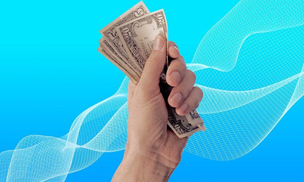 A hand grasping a stack of cash against a vibrant blue background, symbolising financial transactions or wealth