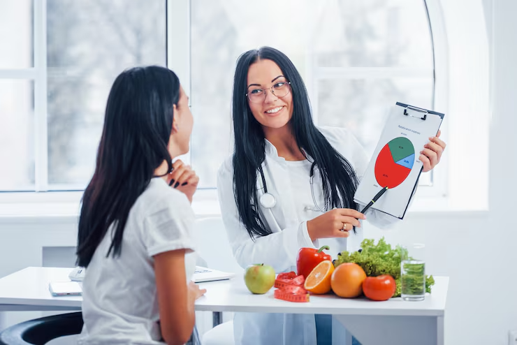 imgi_6_female-nutritionist-with-graph-gives-consultation-patient-indoors-office_146671-29989 Nutritionist explaining diet chart to a woman with fruits and vegetables on the table in a health consultation setting